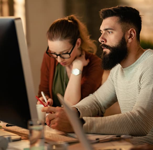 Bearded man and woman in glasses working together at a computer in a dim office